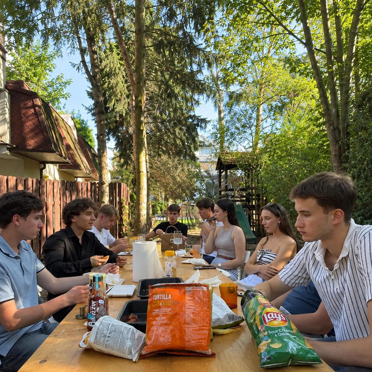 People eating at a long table in the garden under tall trees