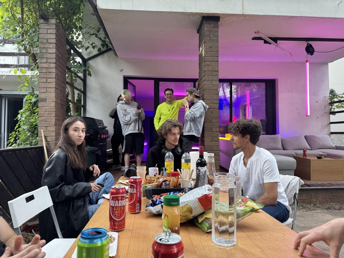 People around a table of food and drinks in front of the house, with pink lights on the porch